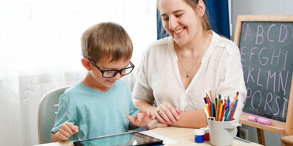 Image of a young student seated at a desk in a classroom, engaging with a tablet while a teacher or TA, sits beside him, smiling.  In the background there is a chalkboard showing the alphabet, and a cup with coloured pencils.