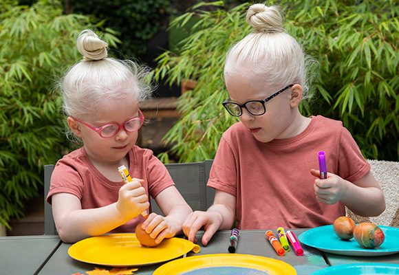 Image showing two young children who are interacting as they decorate eggs, possibly for Easter. The image shows them using coloured pen and plates in bright yellow and turquoise hues.