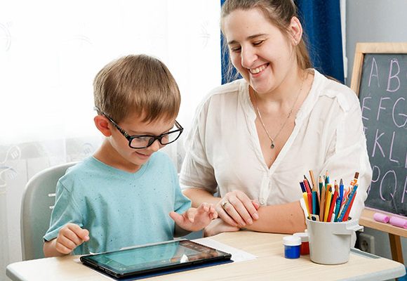 Image of a young student seated at a desk in a classroom, engaging with a tablet while a teacher or TA, sits beside him, smiling.  In the background there is a chalkboard showing the alphabet, and a cup with coloured pencils.