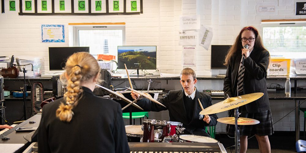 Image of three students in black school uniforms playing drums, keyboard, and singing in a music classroom with computers in the background.