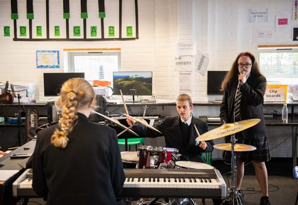 Image of three students in black school uniforms playing drums, keyboard, and singing in a music classroom with computers in the background.