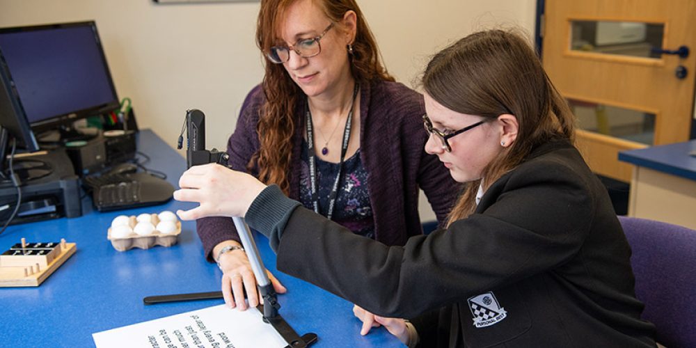 The image depicts a student seated at a desk, engaging with a Pennote while a teacher sits beside them. A computer is visible in the background.