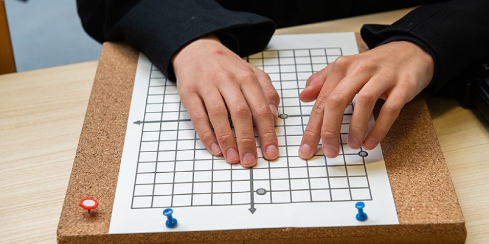 The image shows the hands of a student using a tactile Braille quadrant graph board to plot points in maths