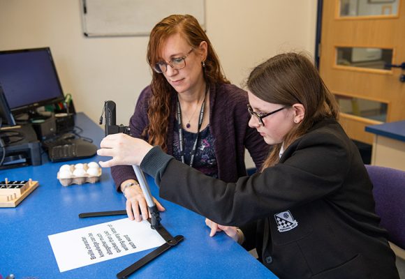 The image depicts a student seated at a desk, engaging with a Pennote while a teacher sits beside them. A computer is visible in the background.