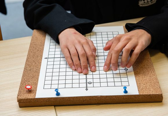 The image shows the hands of a student using a tactile Braille quadrant graph board to plot points in maths