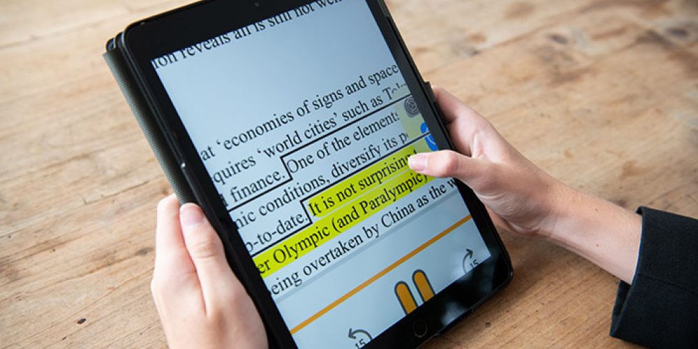 The image shows a person holding a tablet displaying a document with large font, with a section of text highlighted in yellow. The tablet is being used on a wooden surface.