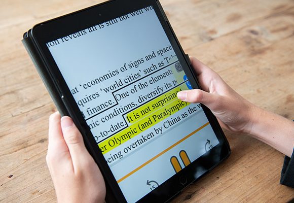 The image shows a person holding a tablet displaying a document with large font, with a section of text highlighted in yellow. The tablet is being used on a wooden surface.